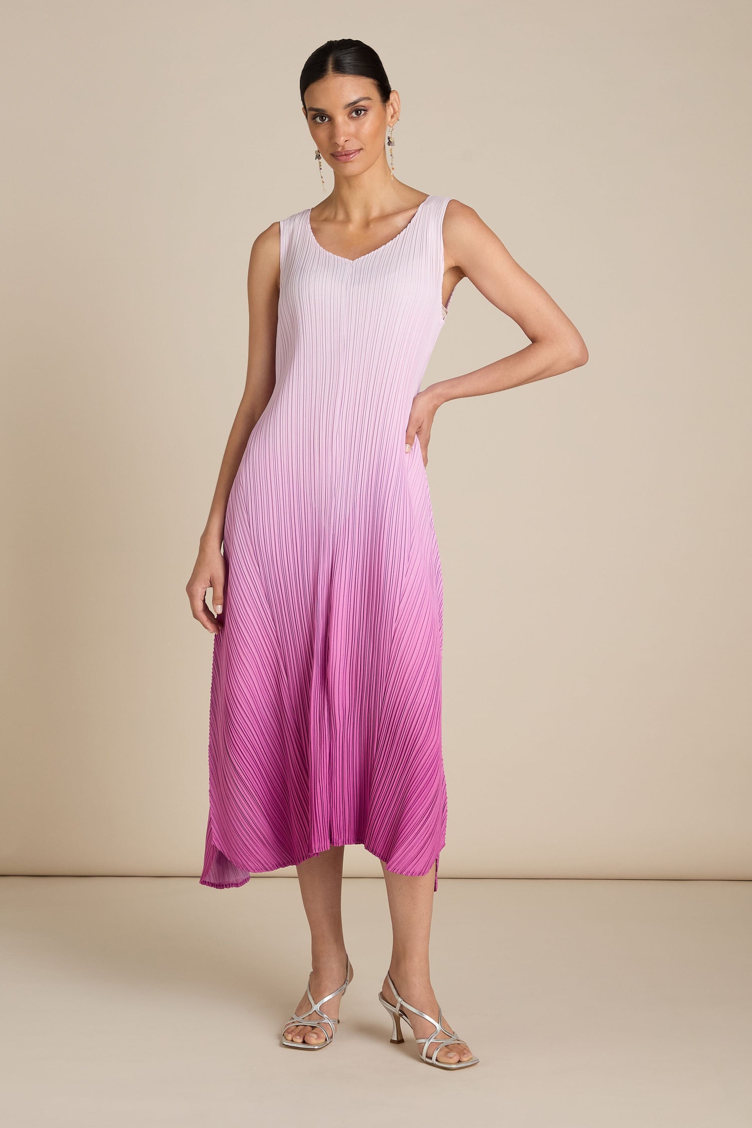 A woman stands confidently in a sleeveless Pleated Dress with an ombre transition from light pink to magenta. The dress features a relaxed silhouette that adds a touch of bohemian flair. She completes her look with metallic-heeled sandals and small hoop earrings.