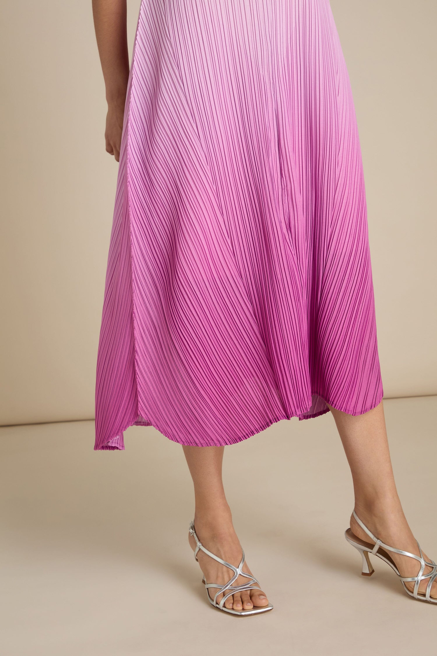 A person is wearing a Pleated Dress in a pink-to-purple ombre with thin straps and silver stilettos adorned with delicate straps, exuding a bohemian flair. The background is beige.