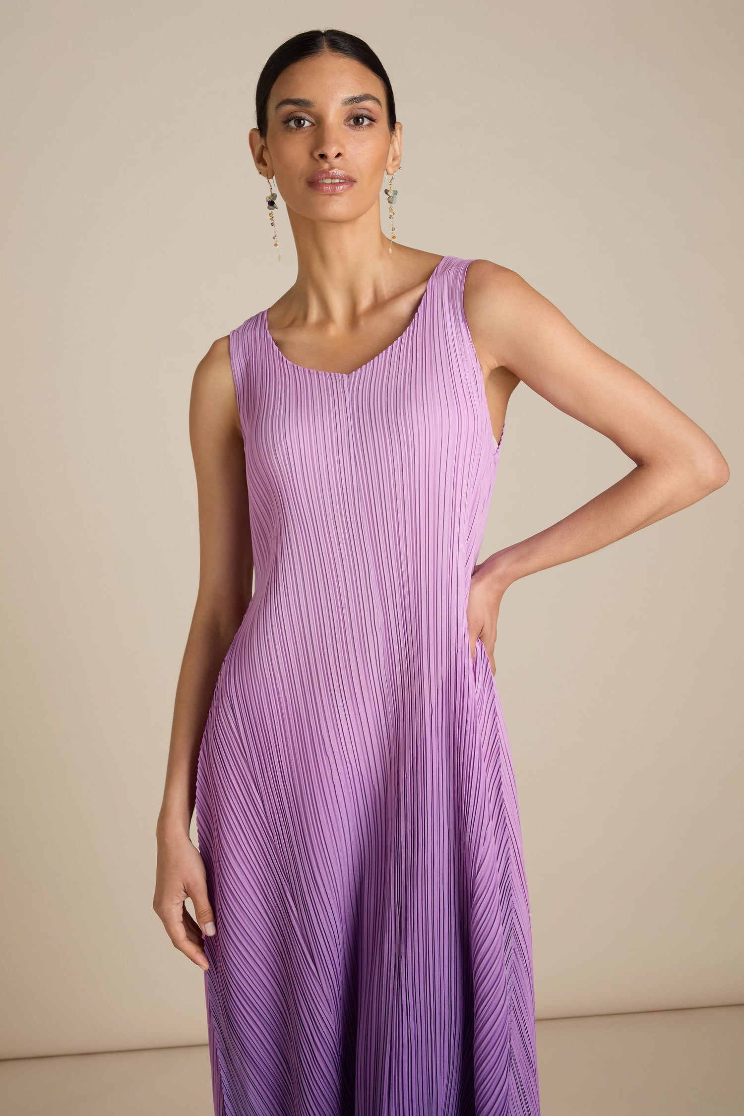 A person stands against a plain background, wearing the Pleated Dress in lavender, its sleeveless design showcasing a relaxed silhouette as one hand rests on their hip.