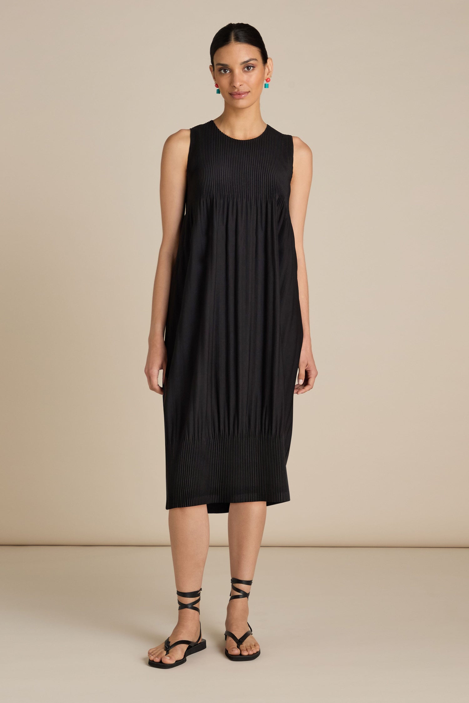 A person stands against a beige background wearing an Alquema Pleated Babydoll Dress in black, accessorized perfectly with statement necklaces and black sandals.