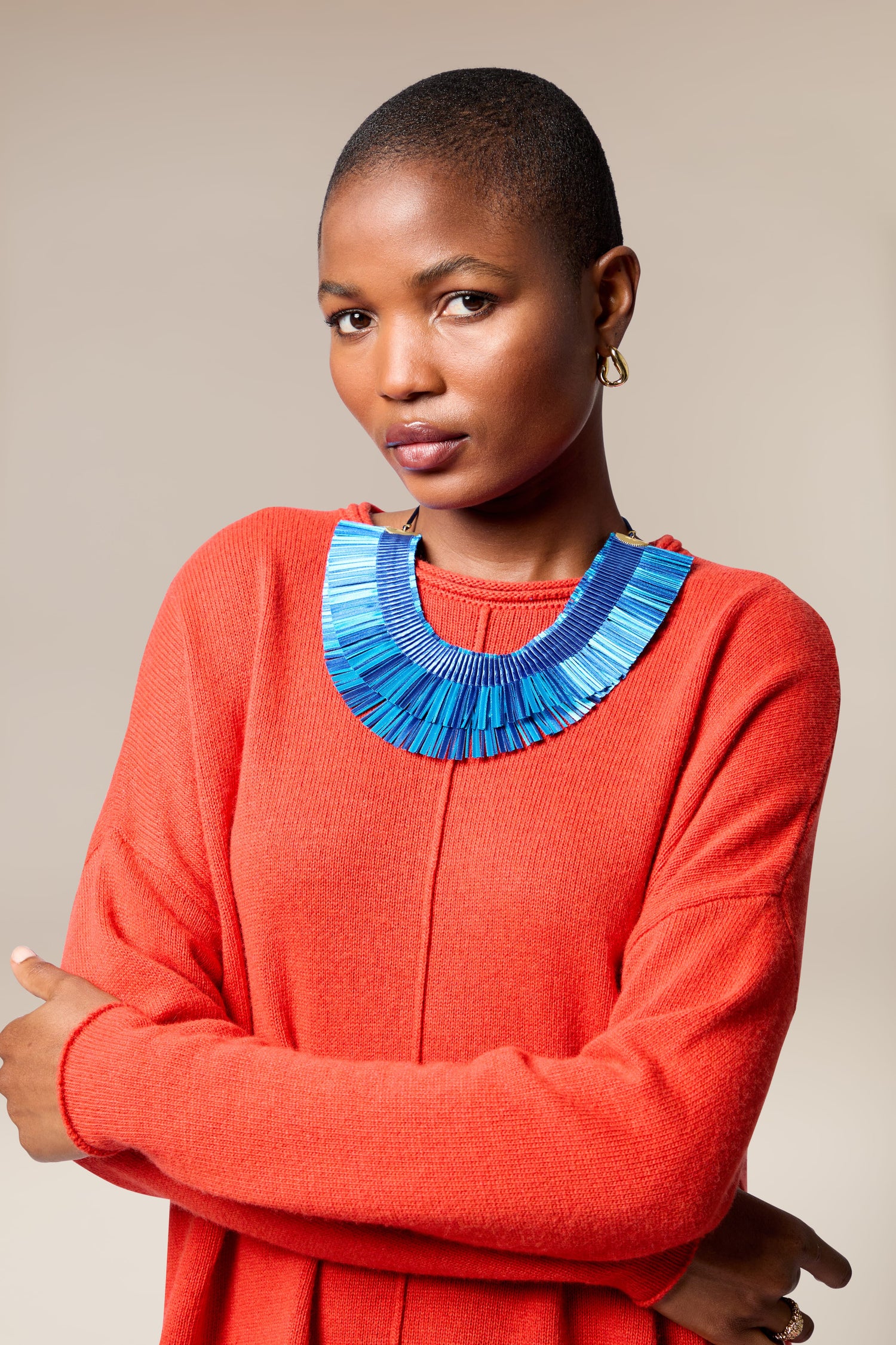 A person with short hair wears a bright orange sweater and the Double Fringe Necklace in bold blue, standing with arms crossed against a neutral background.