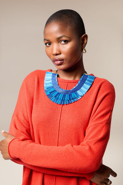 A person with short hair wears a bright orange sweater and the Double Fringe Necklace in bold blue, standing with arms crossed against a neutral background.