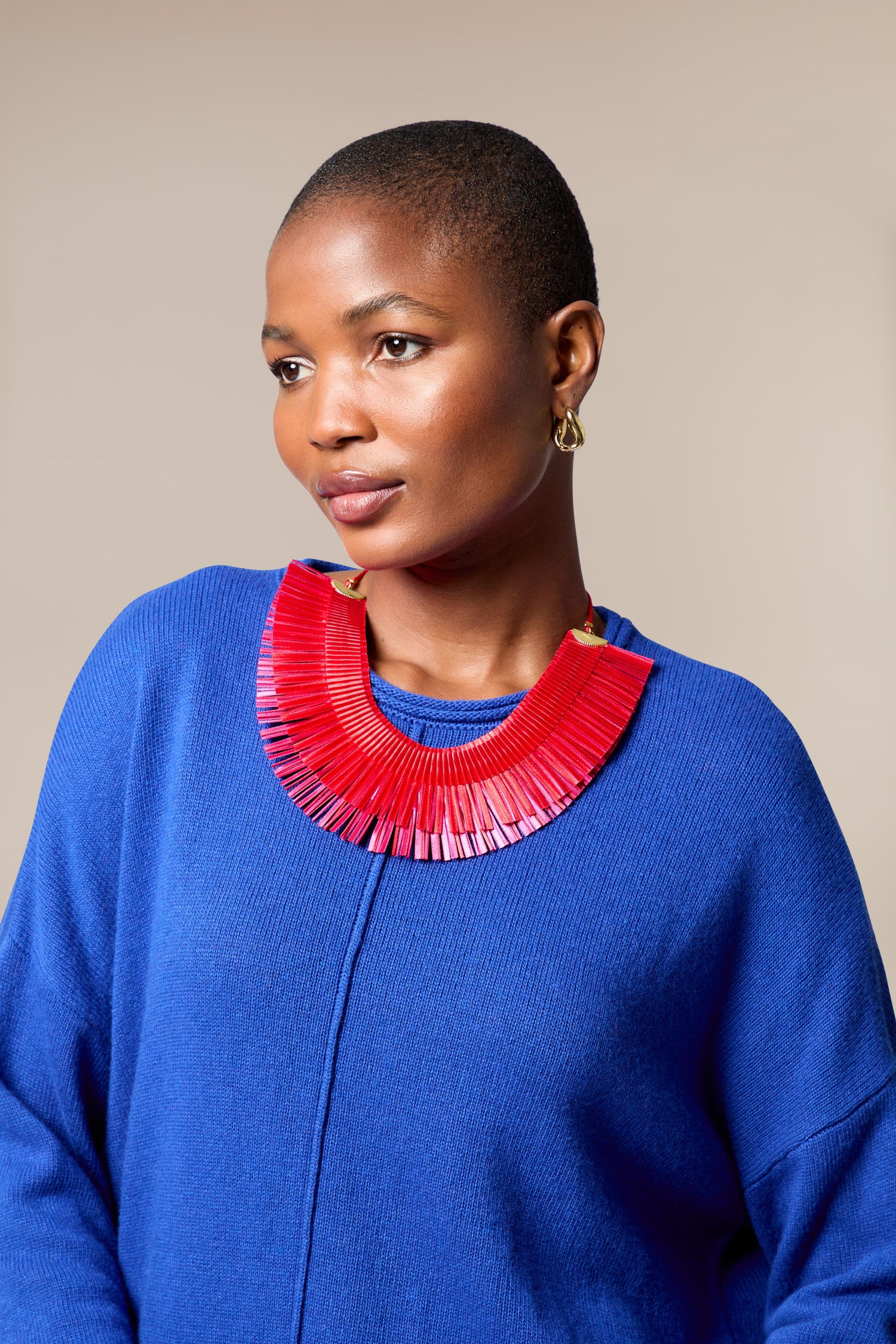 A woman wearing a cobalt blue top, the Double Fringe Necklace in bold red, and gold hoop earrings stands against a neutral background.