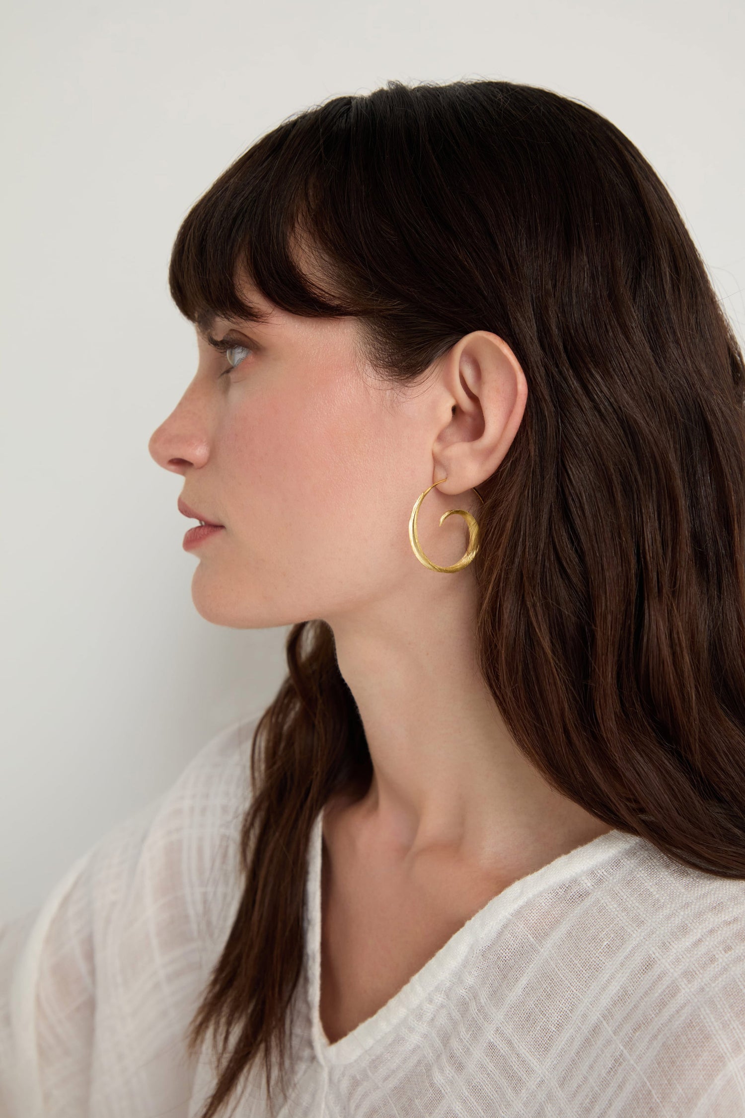 Woman with long brown hair wearing a white textured top and Gold Vermeil Swirl Earrings, facing left against a plain background.