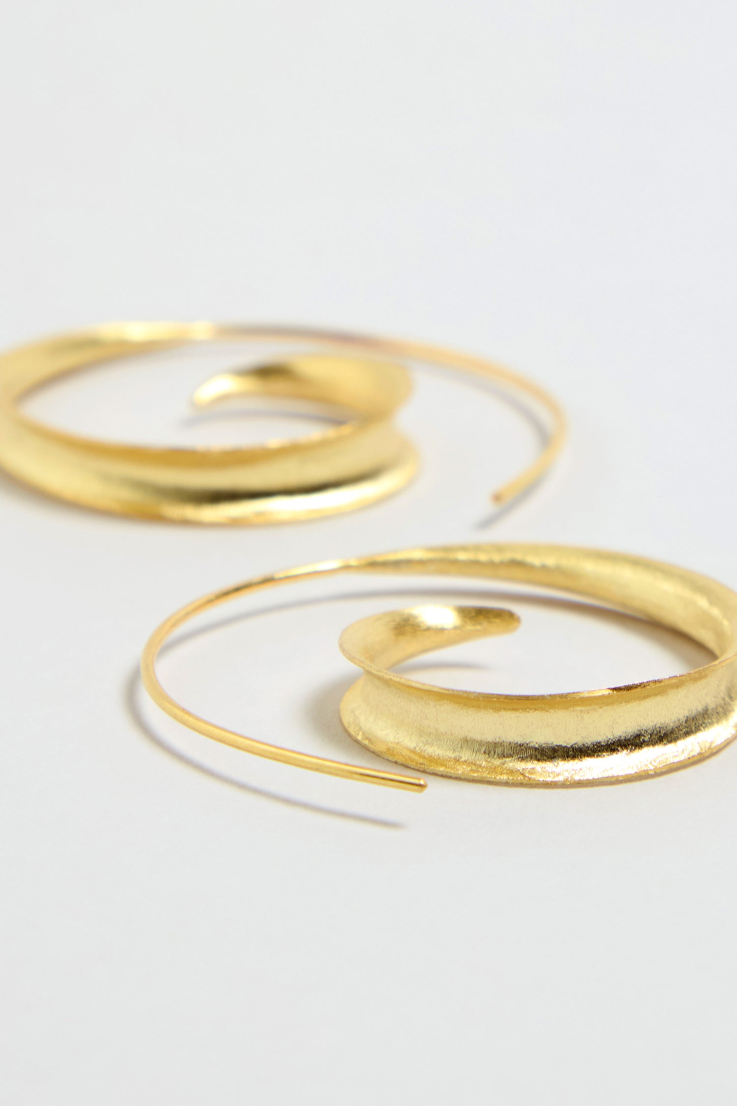 The Gold Vermeil Swirl Earrings rest on a light surface, showcasing sculptural spiral hoops with a modern, elegant design.