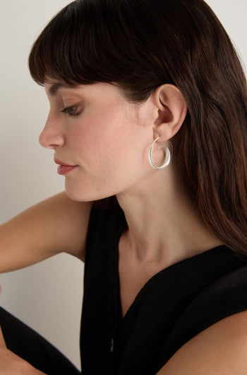 A woman with long brown hair wears the Silver Swirl Earrings, styled with a sleeveless black top and photographed in profile against a plain background.