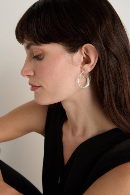 A woman with long brown hair wears the Silver Swirl Earrings, styled with a sleeveless black top and photographed in profile against a plain background.