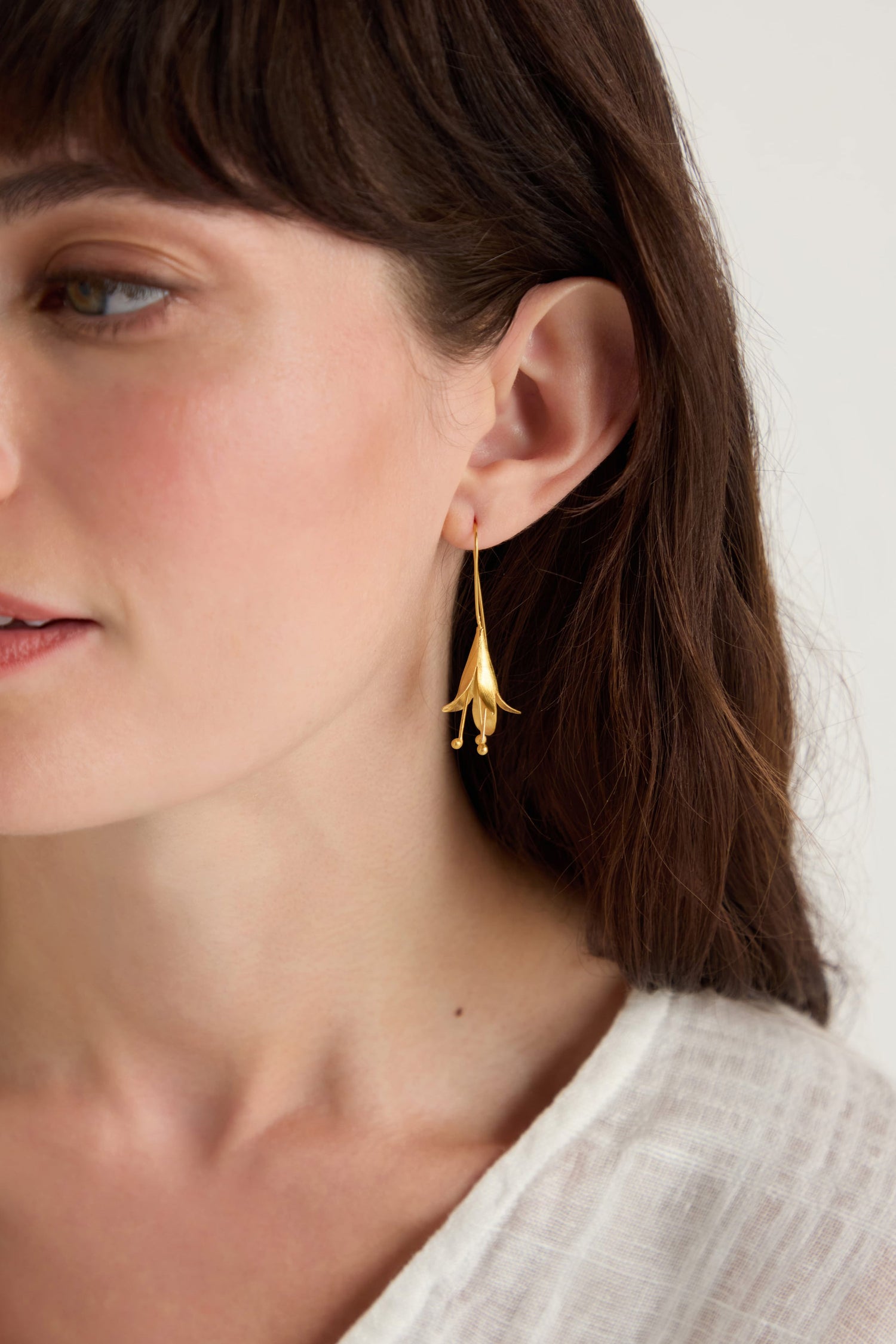 A woman with long brown hair, shown from the neck up and slightly turned to the side, wears Gold Vermeil Flower Earrings and a white textured top.