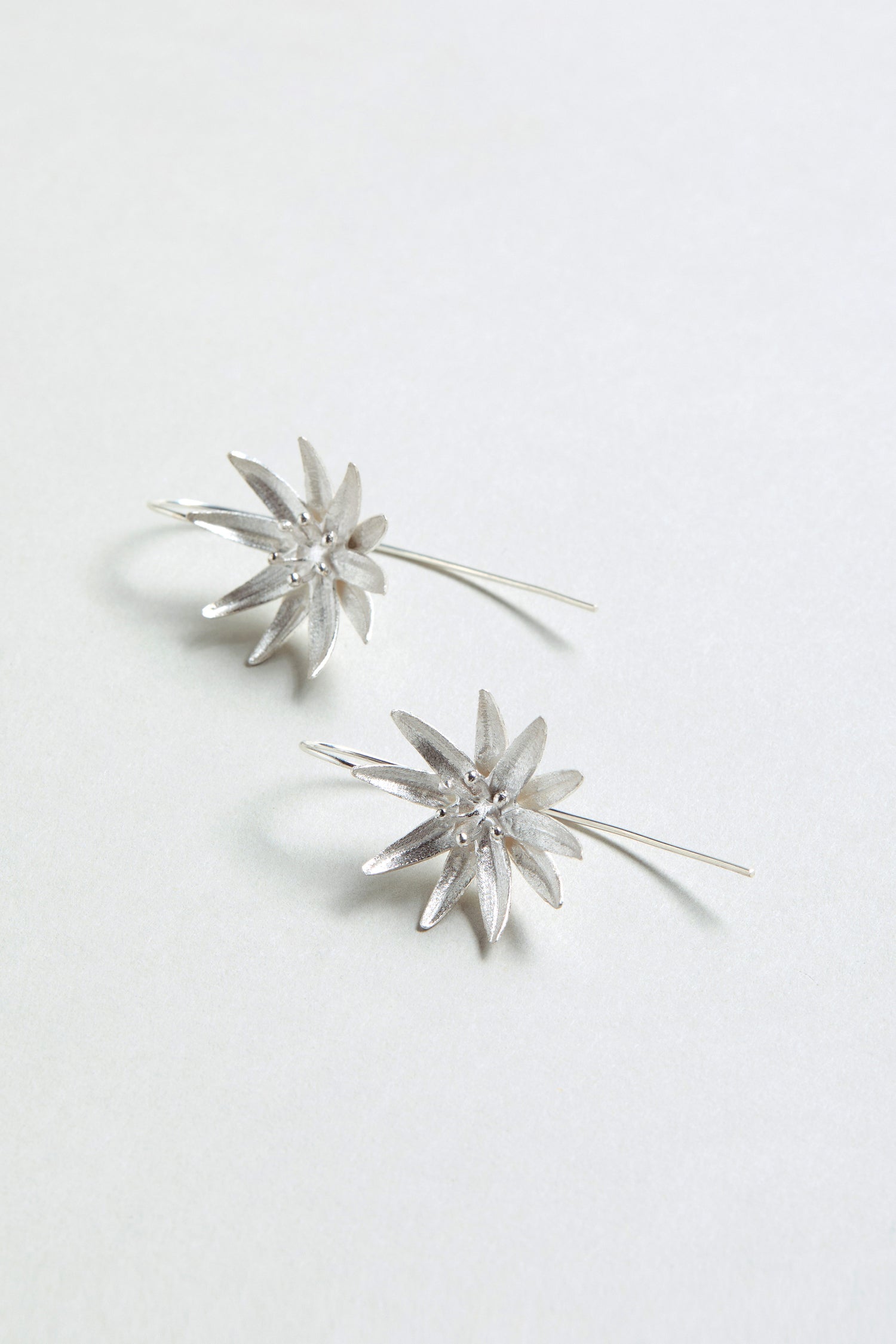 The Silver Starflower Earrings feature sterling silver, star-shaped drops with long, straight hooks, displayed on a light, neutral background—a chic update to classic starflower designs.