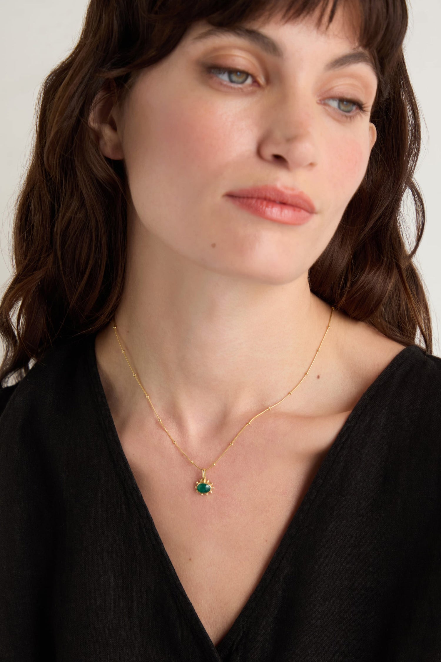 A woman with long brown hair wears a black top and the Gold Vermeil Green Onyx Necklace, featuring a faceted green onyx pendant, as she looks slightly to the side.