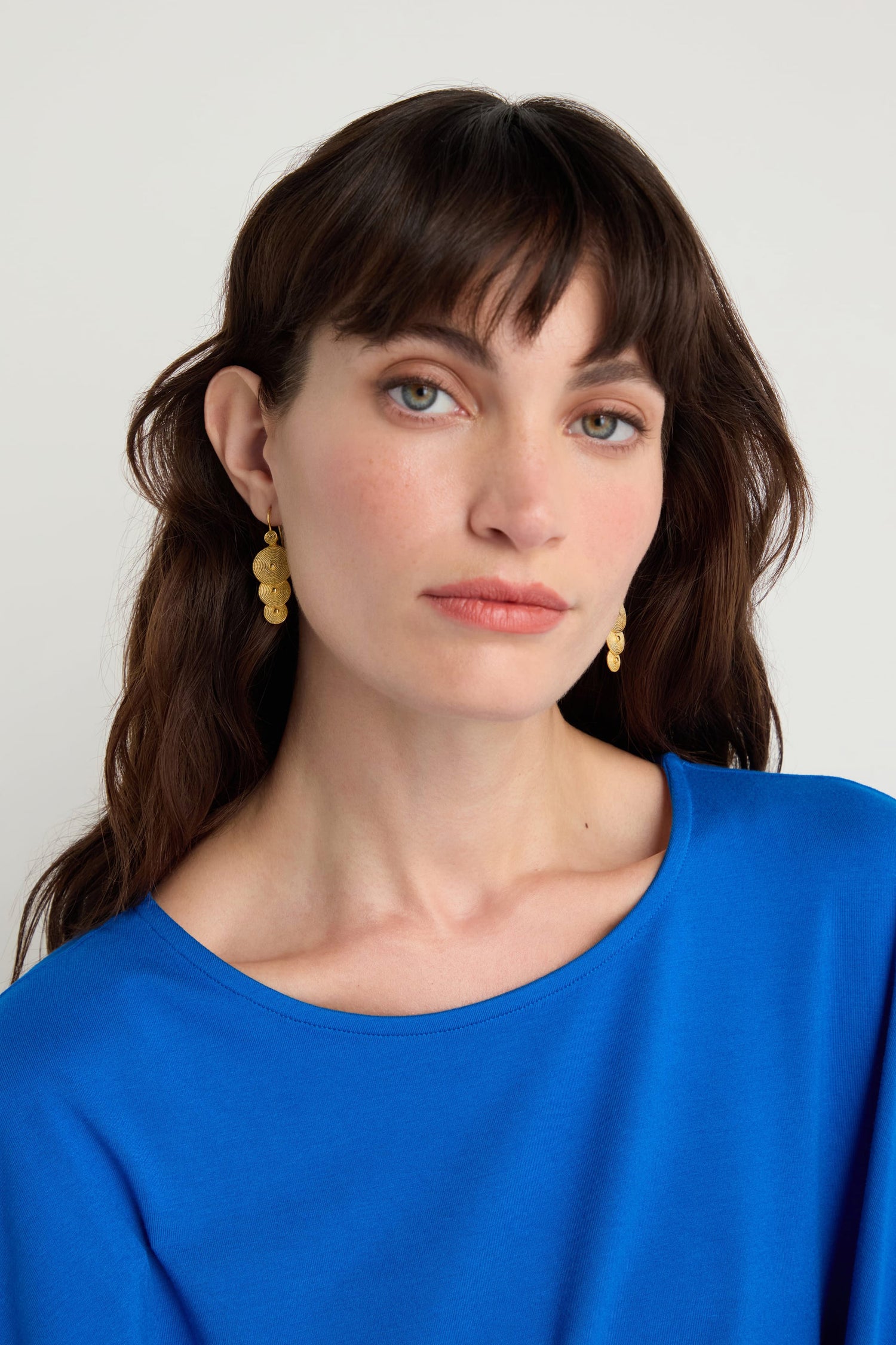 Woman with long brown hair and bangs wearing a bright blue top and Gold Vermeil Orbit Disc Earrings, posed against a plain light background.