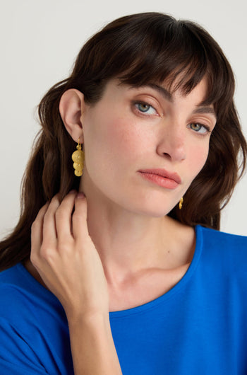 Woman with long brown hair and bangs, wearing a blue shirt and Gold Vermeil Orbit Disc Earrings, looks at the camera with a neutral expression while touching her neck.