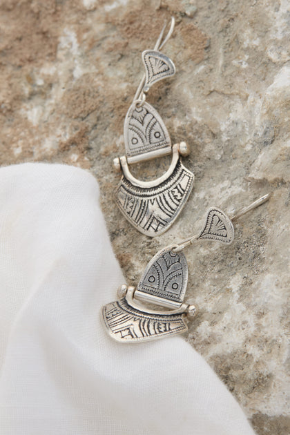 Two exquisite Engraved Silver Tuareg Earrings with intricate designs lie on a textured stone surface next to a white cloth, showcasing the elegance of any jewellery collection.