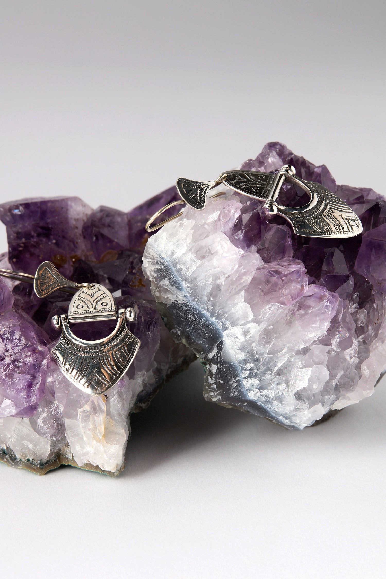 The Engraved Silver Tuareg Earrings, part of a unique jewellery collection, are elegantly displayed on purple amethyst crystal clusters against a gray background.