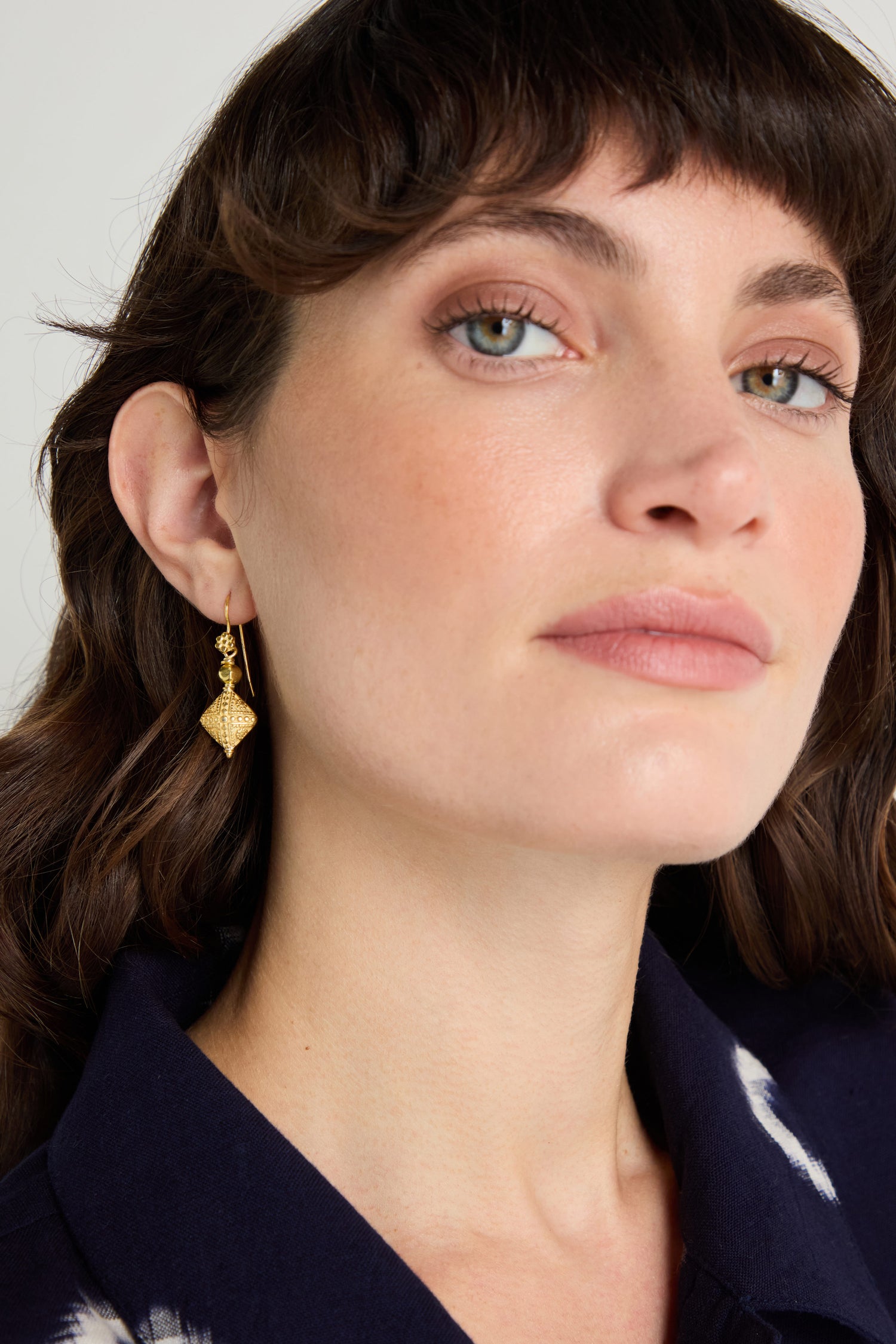 A woman with wavy brown hair wears a navy collared top and Gold Vermeil Faceted Drop Earrings, looking slightly off camera.