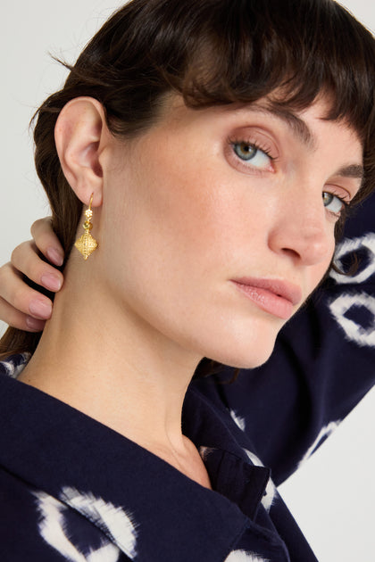 A woman with short dark hair wears a blue patterned top and the Gold Vermeil Faceted Drop Earrings, tilting her head and touching her neck as she looks at the camera.
