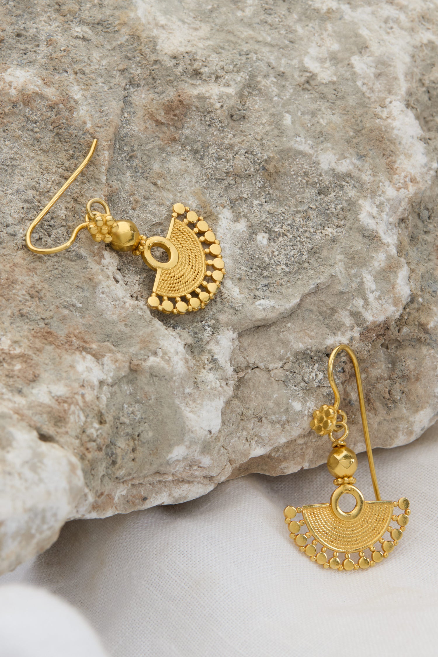 Two intricately designed 18ct Gold Vermeil Mini Fan Earrings are placed on a rough stone surface. One earring dangles from the stone, while the other lies flat beside it, exuding bohemian elegance.