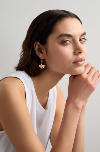 A person with short dark hair in a white sleeveless top rests their chin on their hands. Their Gold Vermeil Mini Fan Earrings add a touch of elegance to their look, being part of an exquisite jewellery collection.