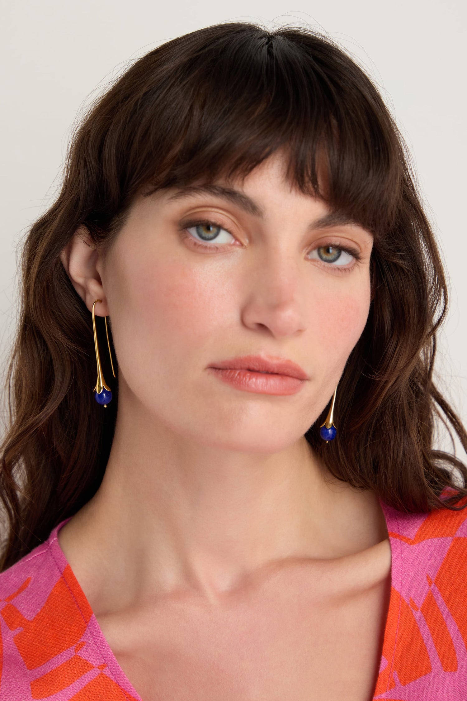 A woman with long brown hair and bangs wears Gold Vermeil Lapis Drop Earrings and a pink patterned top, looking at the camera with a neutral expression.