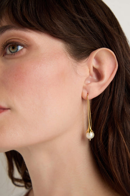 Close-up of a woman wearing Gold Vermeil Pearl Drop Earrings on her left ear, featuring a dangling pearl, set against a neutral background.