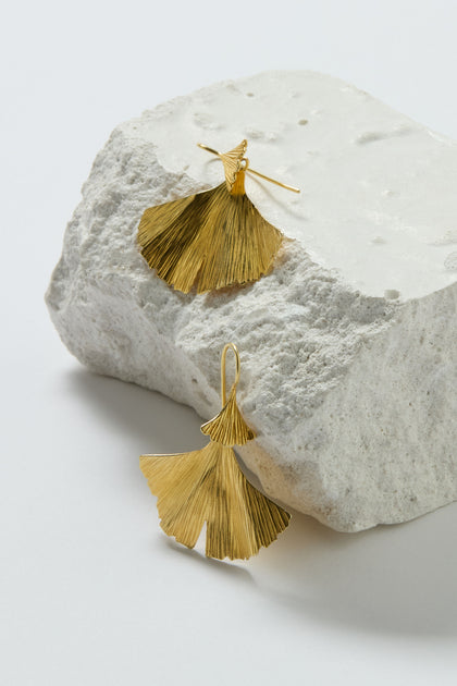 The Gold Vermeil Ginkgo Leaf Earrings, inspired by nature, are shown on and in front of a textured white stone with a plain background.