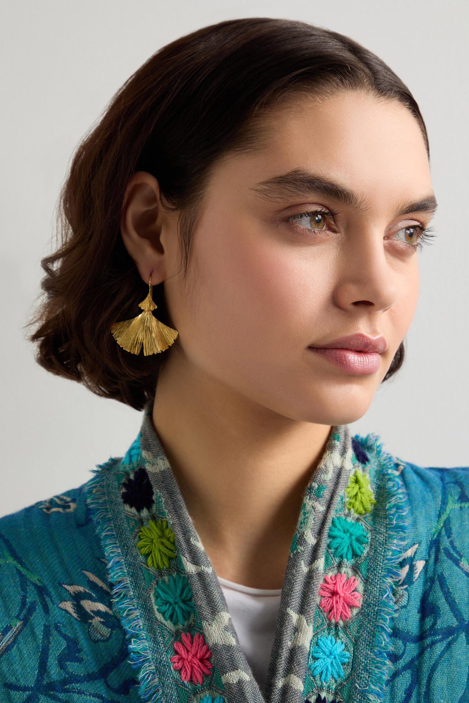 A person with short hair wears Gold Vermeil Ginkgo Leaf Earrings and a colorful embroidered blue jacket, looking to the side.