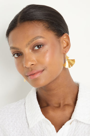 Woman with smooth, pulled-back hair wears a white textured top and Gold Vermeil Ginkgo Leaf Earrings, looking slightly to the side and smiling subtly against a plain background.