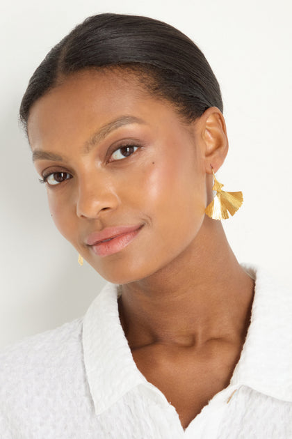 Woman with smooth, pulled-back hair wears a white textured top and Gold Vermeil Ginkgo Leaf Earrings, looking slightly to the side and smiling subtly against a plain background.