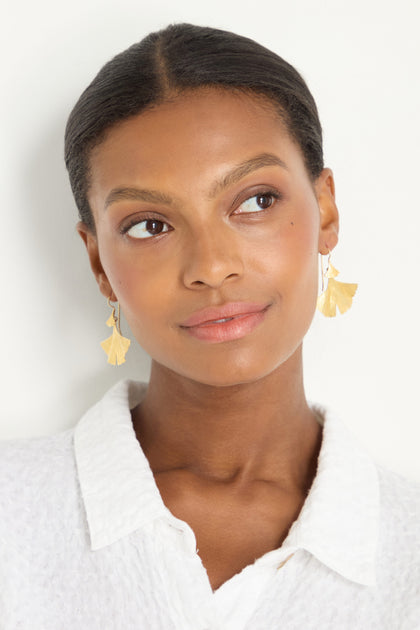 A woman with smooth dark skin and pulled-back hair wears Gold Vermeil Ginkgo Leaf Earrings and a white collared shirt, gazing to the side against a plain background.