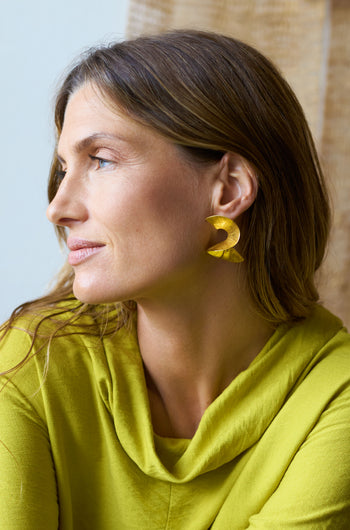 A woman with long brown hair wearing a green top looks to the side, featuring large Gold Vermeil Twist Earrings that catch the light beautifully.