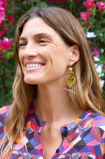 A smiling woman with long hair wears Gold Vermeil Sumba Earrings, enhancing her artisanal look. Her colorful geometric-patterned blouse complements the intricate filigree as she stands gracefully before pink flowers.