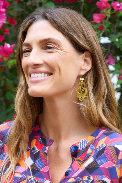 A smiling woman with long hair wears Gold Vermeil Sumba Earrings, enhancing her artisanal look. Her colorful geometric-patterned blouse complements the intricate filigree as she stands gracefully before pink flowers.