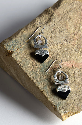 The Silver and Black Onyx Tuareg Earrings, featuring intricate designs, are elegantly displayed on a textured stone surface.