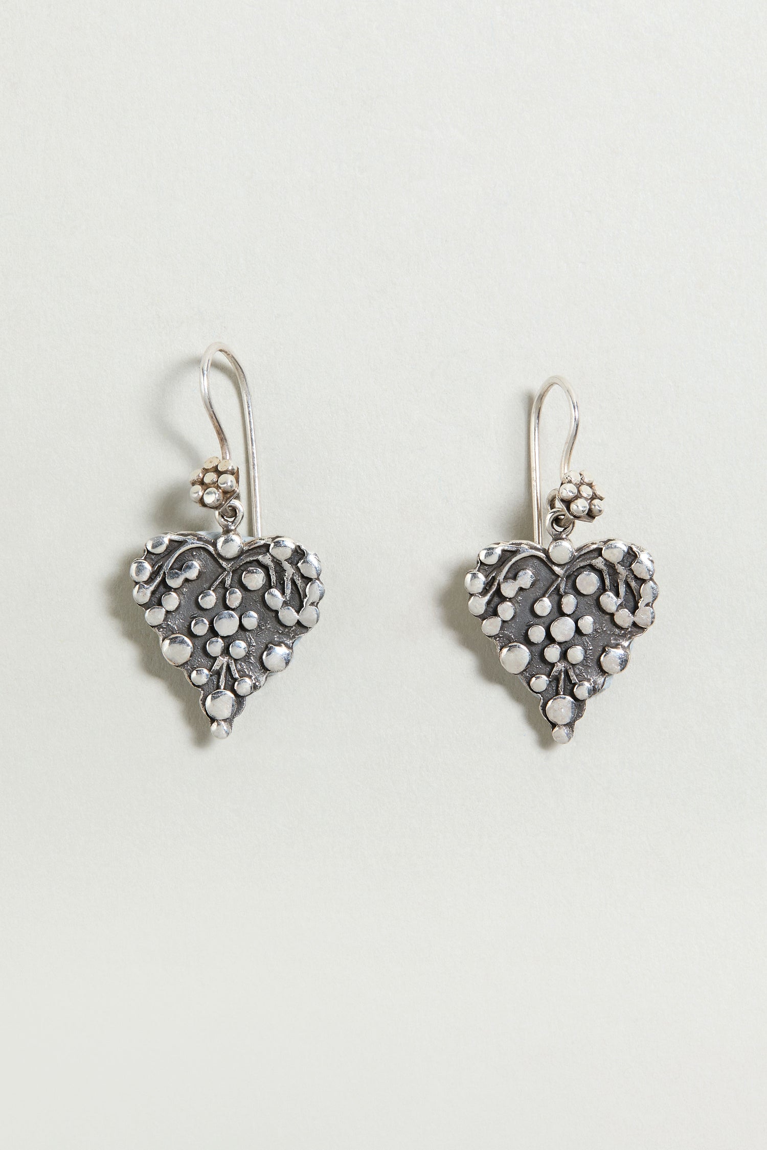 Add a delicate touch to any look with the Silver Heart Earrings—sterling silver drop earrings featuring heart shapes with floral and dot details, perfect against a light background.