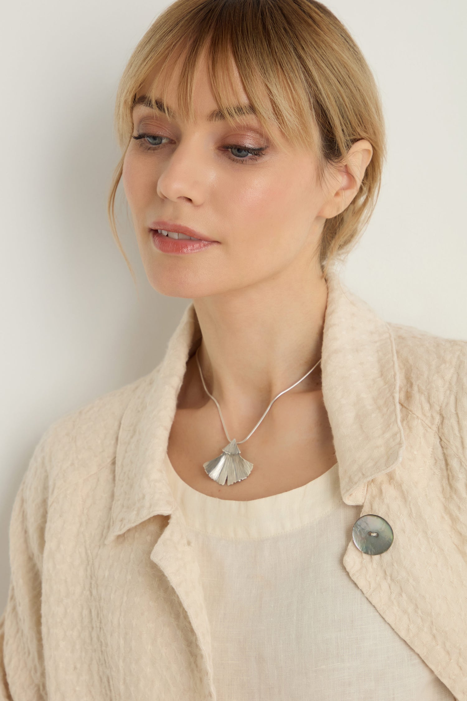A woman with blonde hair wears a textured cream jacket, an off-white top, and the Silver Ginkgo Leaf Pendant, looking down slightly.