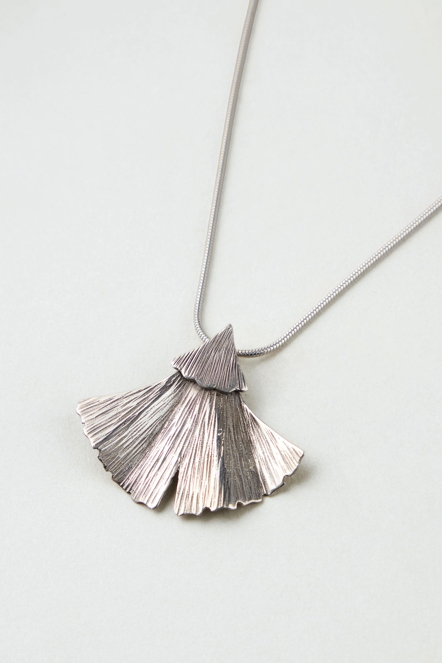 The Silver Ginkgo Leaf Pendant features a textured, fan-shaped design on a thin, smooth chain and is displayed against a light gray background.