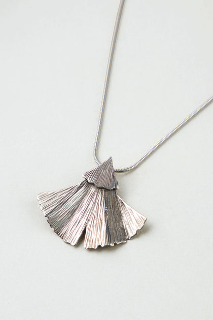 The Silver Ginkgo Leaf Pendant features a textured, fan-shaped design on a thin, smooth chain and is displayed against a light gray background.