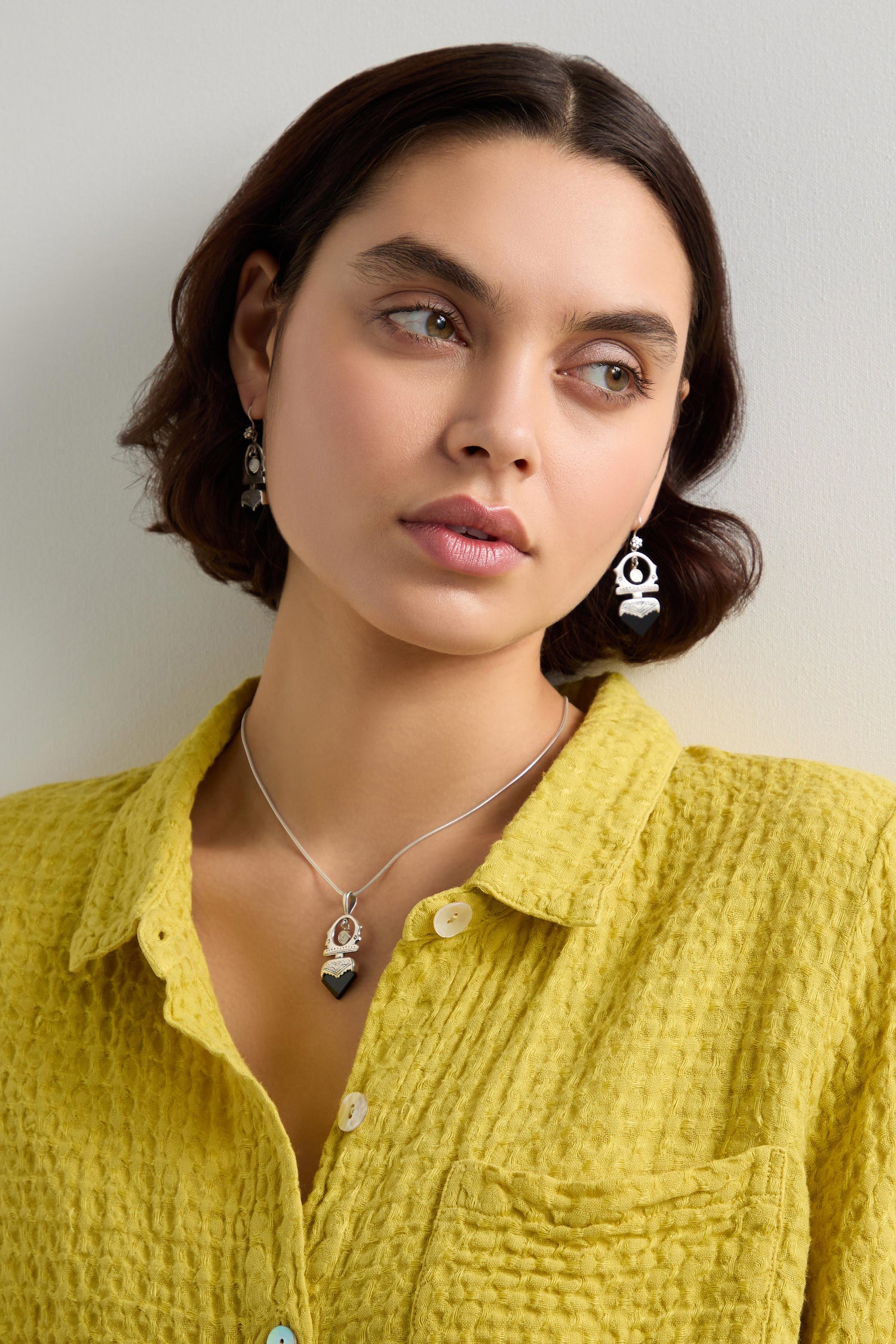 A woman with short dark hair wears a textured yellow shirt, black and silver earrings, and a handcrafted necklace showcasing a Tuareg Silver and Black Onyx Pendant, as she looks to the side against a plain background.