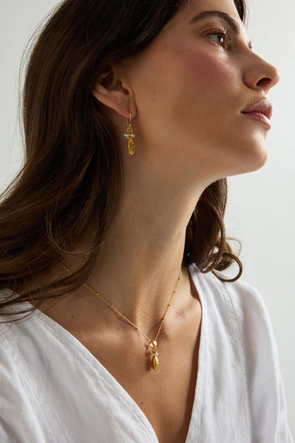 A woman wearing a white V-neck top, gold earrings, and the Gold Vermeil Pearl Drop Necklace with a freshwater pearl pendant, gazes slightly upward against a plain background.