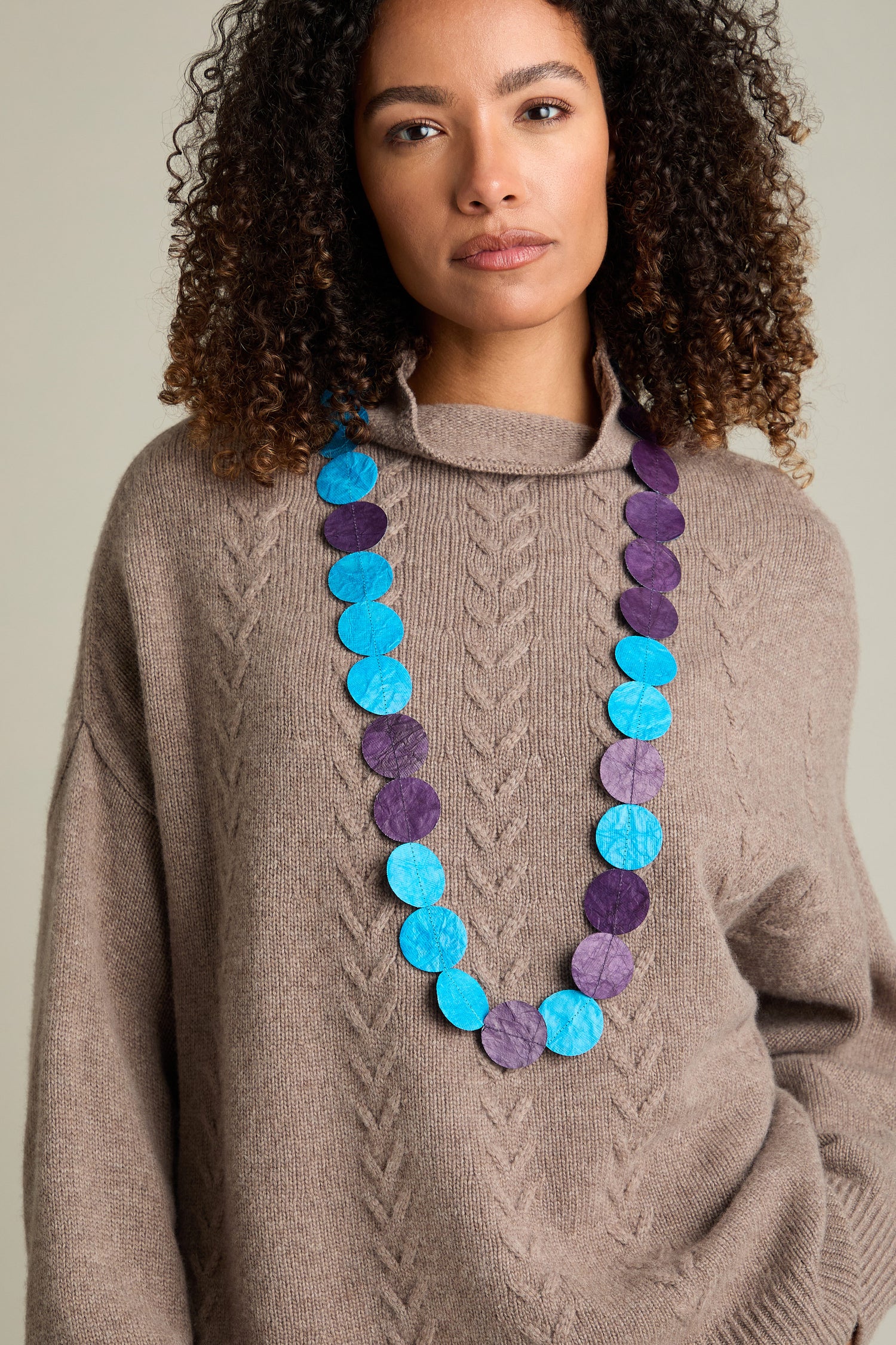 A person with curly hair wearing a beige sweater and the Dotty Necklace, featuring circular paper beads in blue and purple elements, stands against a plain background.