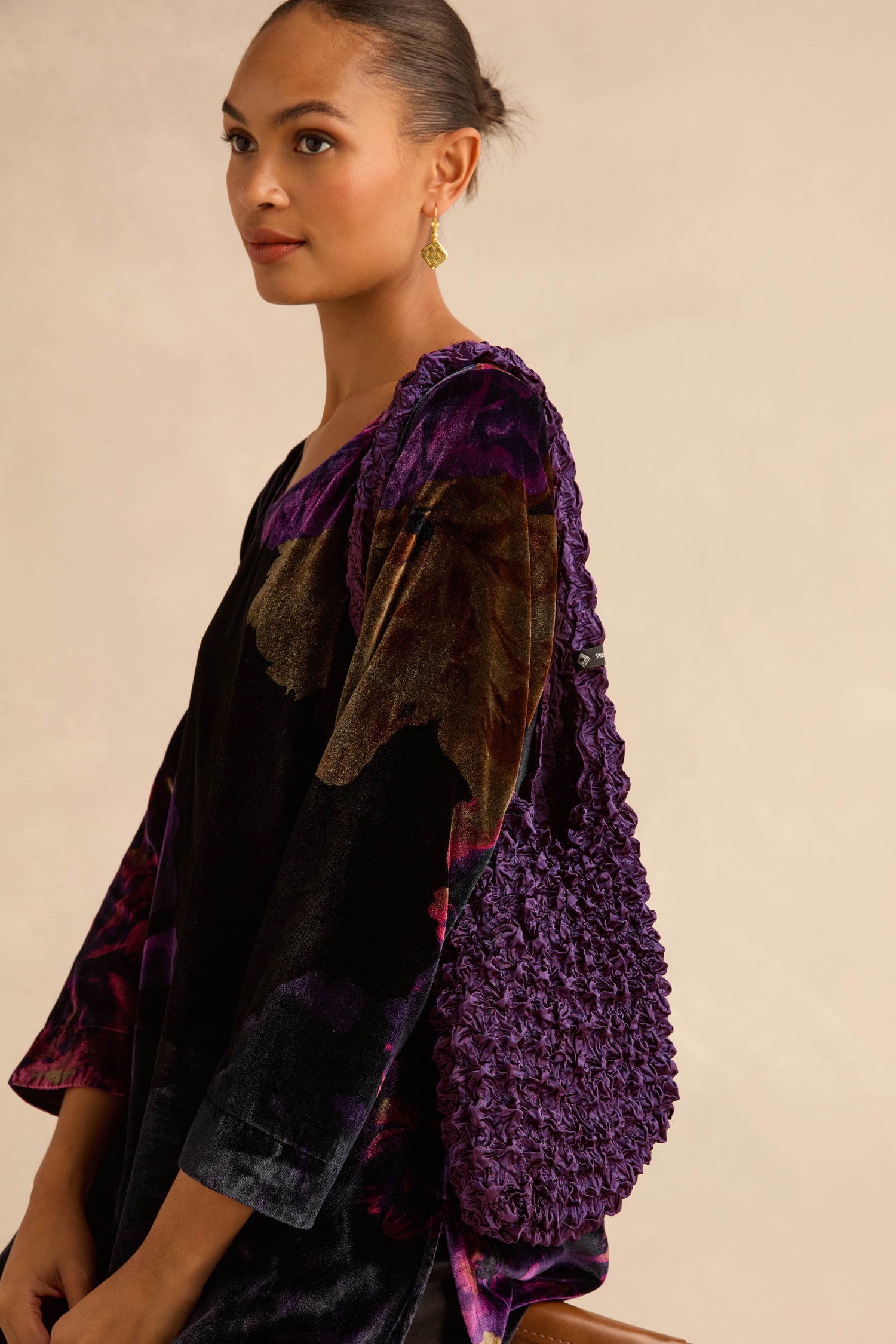 A person wearing a dark purple floral tunic carries an Aubergine Shibori Pleated Bag, a textured, deep purple pleated accessory.