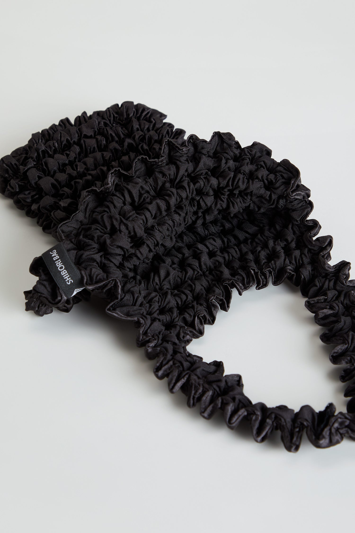 Black Shibori Pleated bag featuring pleats, a gathered handle, a small “SHIBORI BAG” label, and an expandable tote design.
