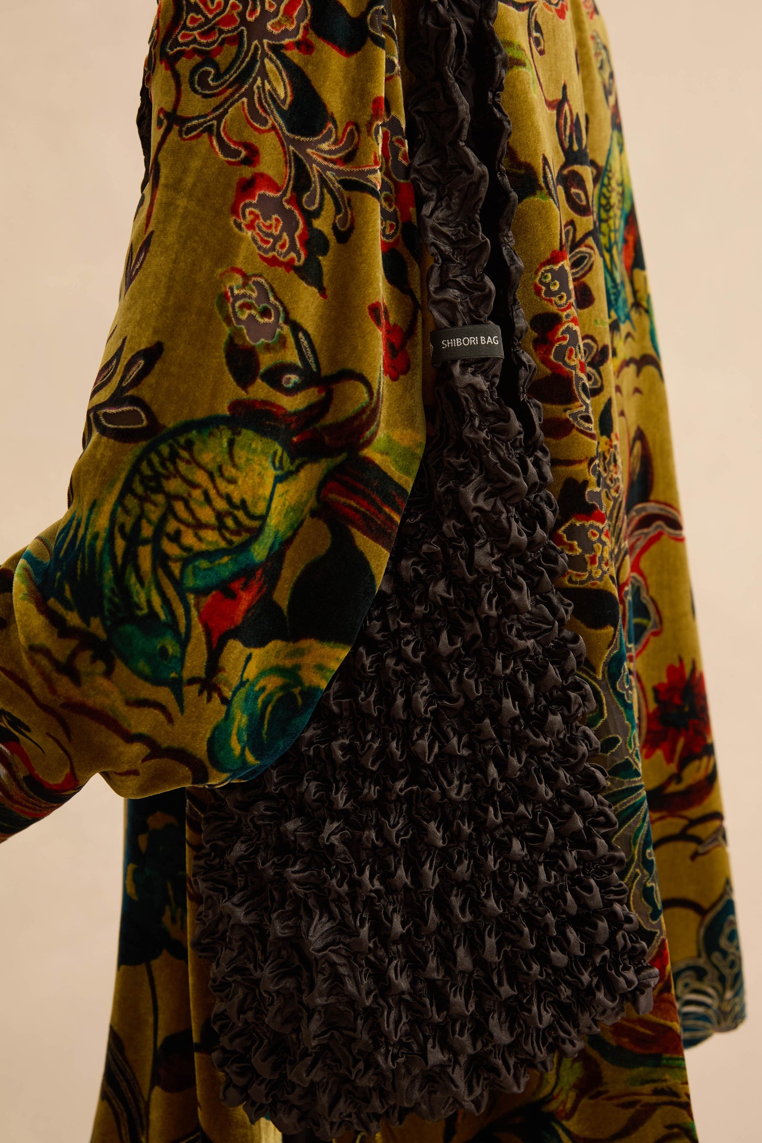 A Black Shibori Pleated Bag worn over a shoulder against a vibrant, multi-colored floral-patterned garment.