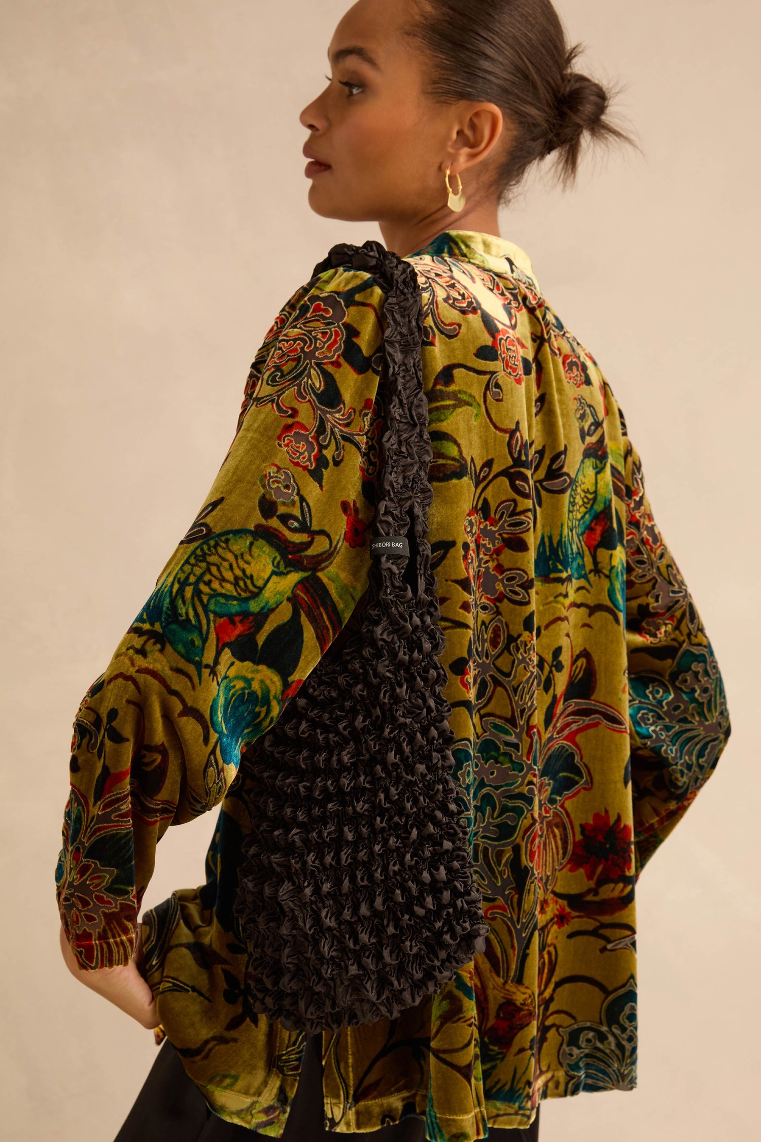 A person wears a Black Shibori Pleated Bag over a floral patterned velvet top.