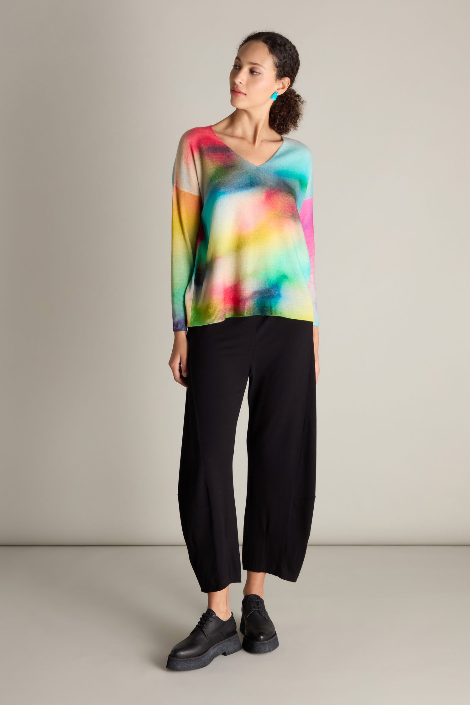 A person standing and gazing sideways, dressed in a colorful V-Neck Cashmere Jumper featuring a rainbow watercolor print, paired with black wide-leg pants and black shoes.