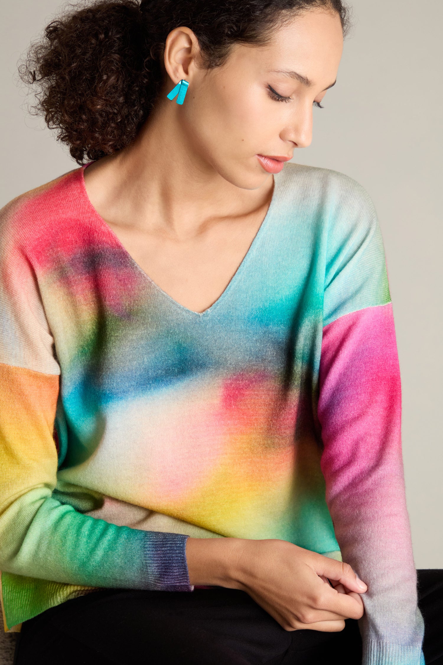 A woman with curly hair is wearing a V-Neck Cashmere Jumper in a vibrant rainbow watercolor print and blue earrings, looking downwards.