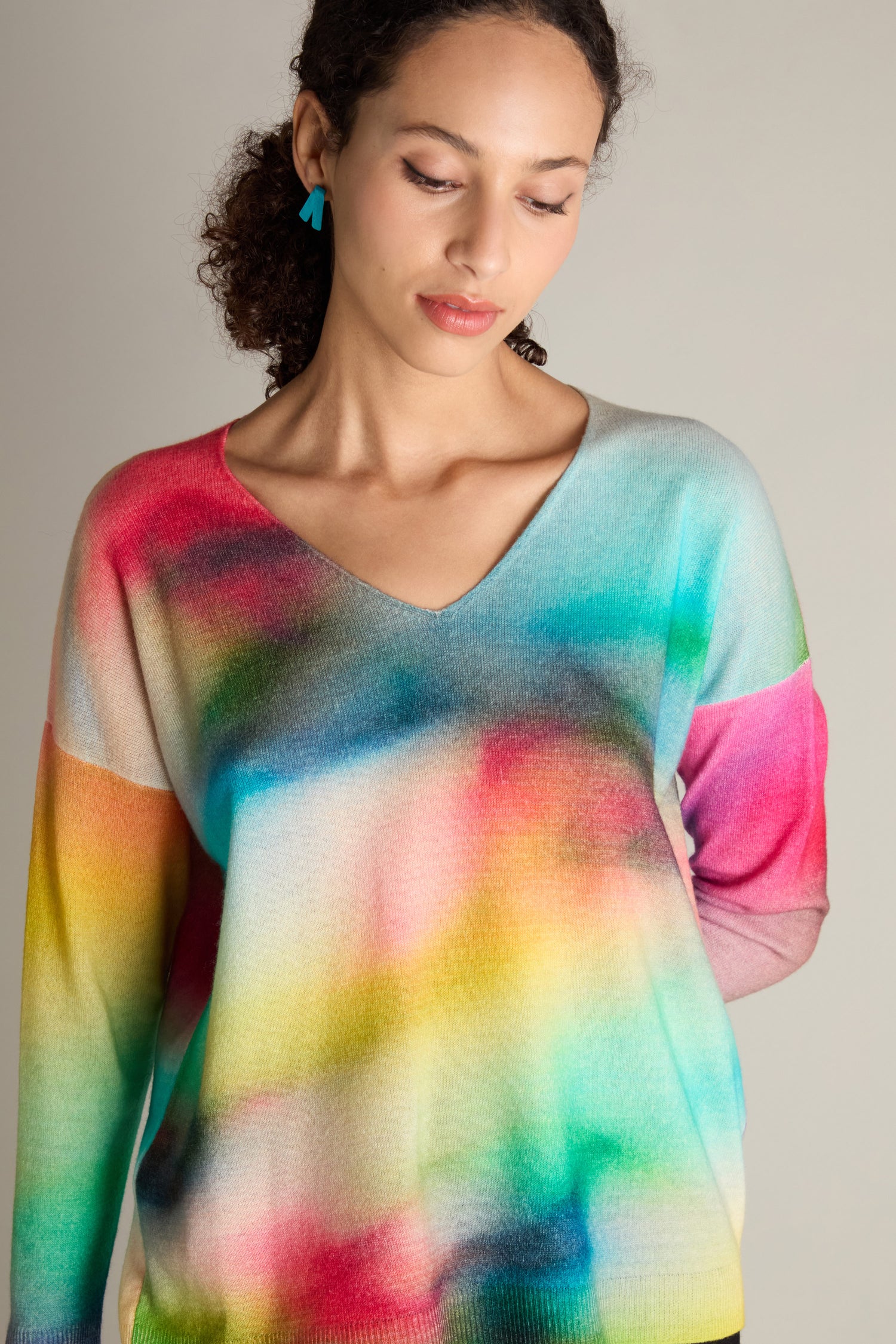 A woman wearing a V-Neck Cashmere Jumper adorned with a rainbow watercolour print gazes downward with a neutral expression.