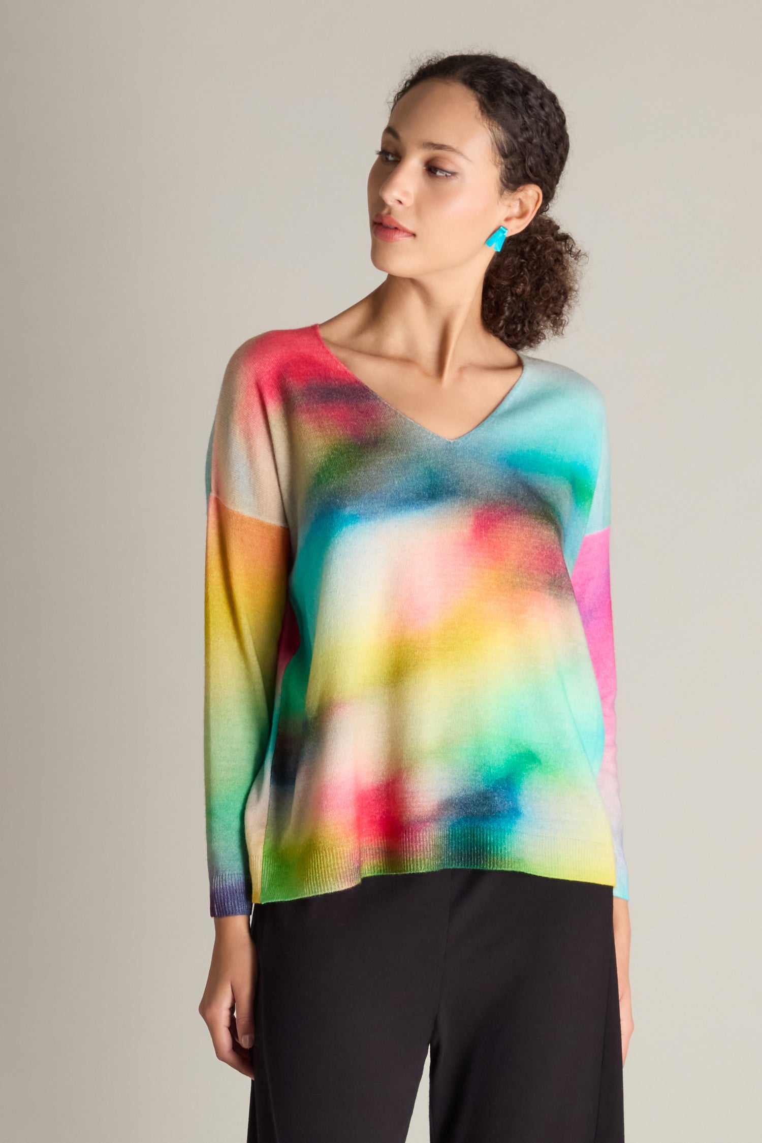 A woman wears a V-Neck Cashmere Jumper featuring a colorful, gradient rainbow watercolor print and black pants, standing against a plain background, looking to her right.