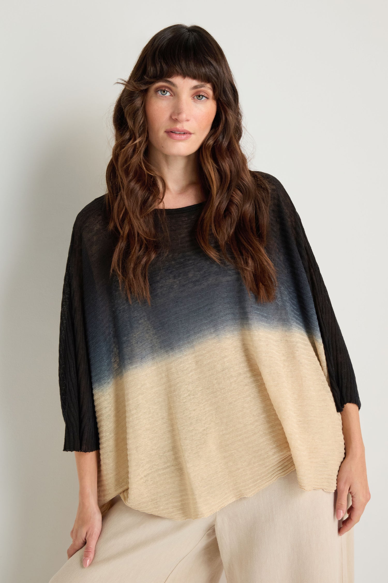 A woman with long wavy brown hair models the Dip Dye Knit Easy Top in black, blue, and beige tones with light pants, standing against a plain background.