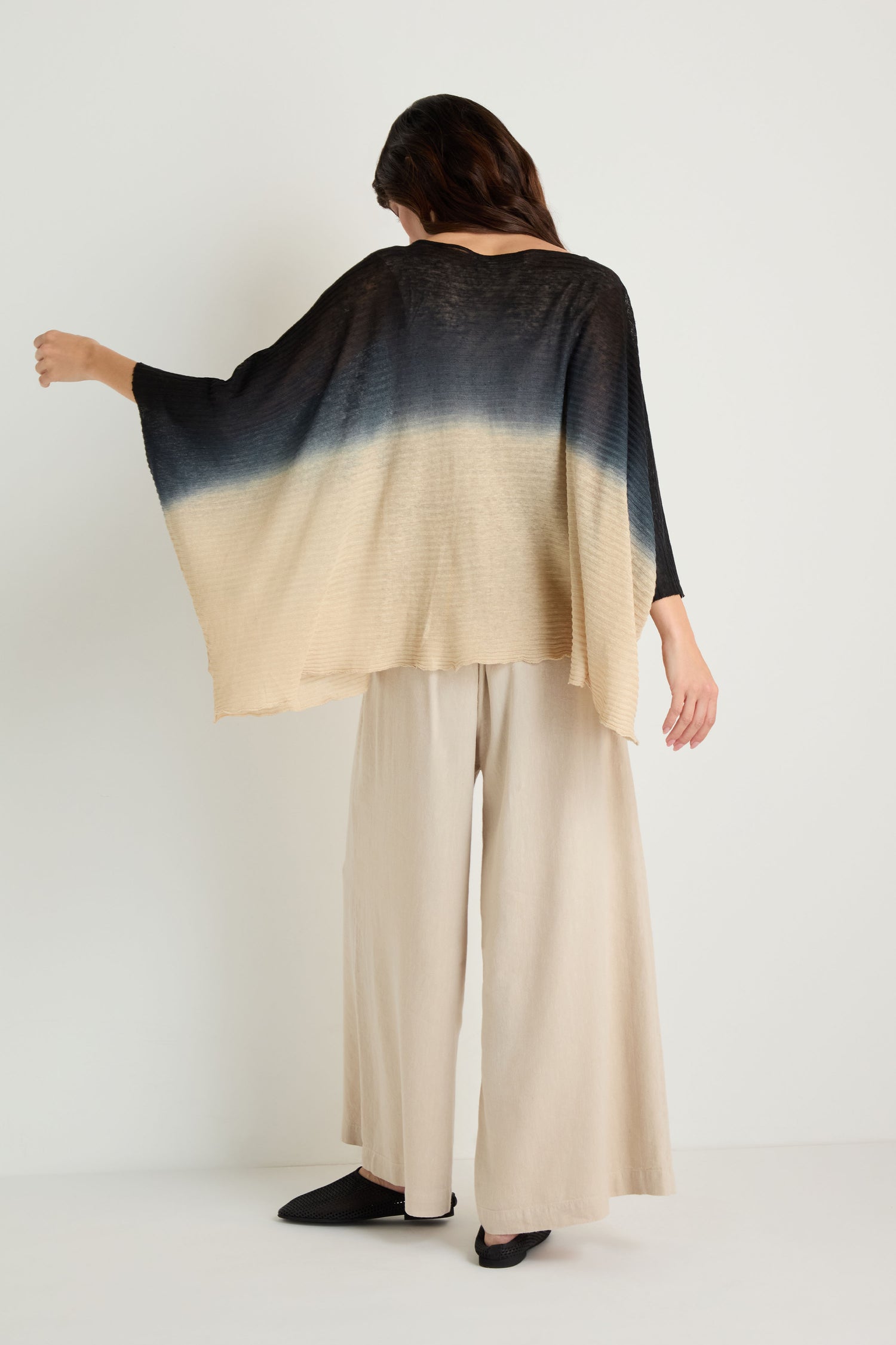 A person stands facing away, wearing wide-leg beige pants and the Dip Dye Knit Easy Top, which features a loose fit and a transition from black to beige.